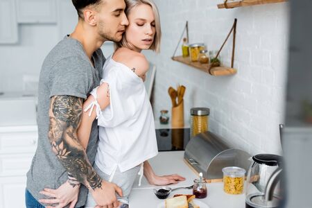 Side view of tattooed man touching beautiful blonde girlfriend near breakfast in kitchenの写真素材