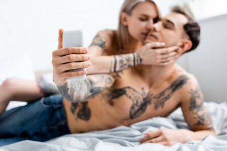 Selective focus of girl kissing tattooed boyfriend taking selfie with smartphone on bedの写真素材