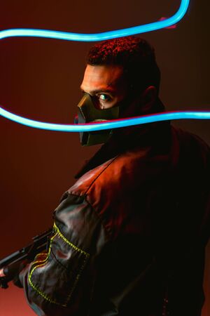 selective focus of bi-racial cyberpunk player in protective mask holding gun near blue neon lighting on blackの写真素材
