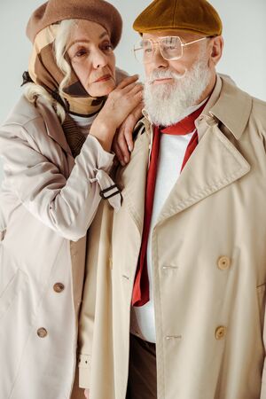 Beautiful senior woman embracing and looking at elegant man in trench coat isolated on grey