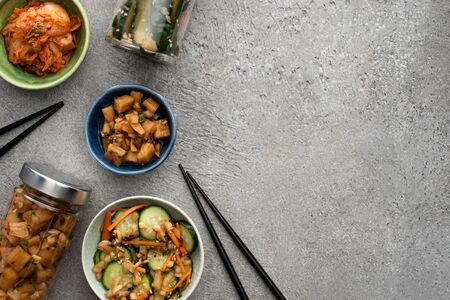top view of tasty kimchi in bowls and jars near chopsticks on concrete surface