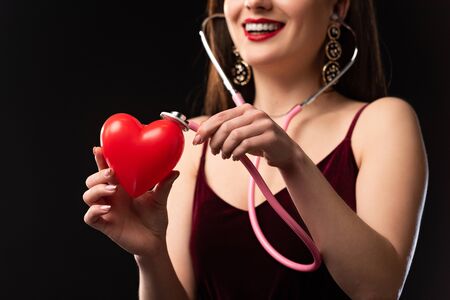 cropped view of smiling woman in dress holding heart-shaped model and stethoscope isolated on black