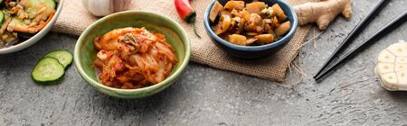 panoramic shot of bowls with kimchi near chopsticks, ginger, chili pepper, sliced cucumber and garlic on concrete surfaceの写真素材