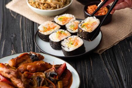 cropped view of woman holding chopsticks with rice roll near korean side dishes