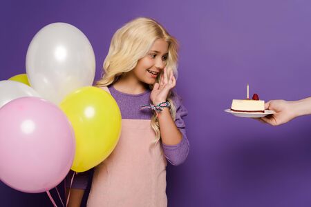 cropped view of woman giving plate with birthday cake to smiling kid with balloons on purple backgroundの写真素材