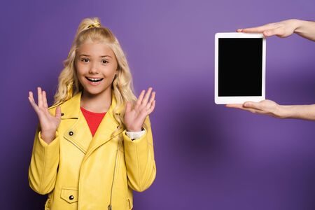 cropped view of man giving digital tablet to surprised kid on purple background