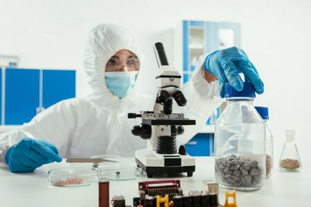 engineer in hazmat suit holding jar with gravel near microscope and computer motherboardの写真素材
