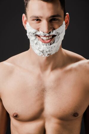 cheerful naked man with shaving foam on face isolated on blackの写真素材