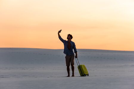 silhouette of man walking on beach with travel bag and smartphone at sunsetの写真素材