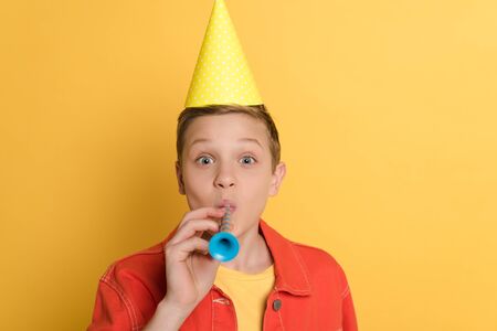 kid in party cap blowing in party horn on yellow backgroundの写真素材