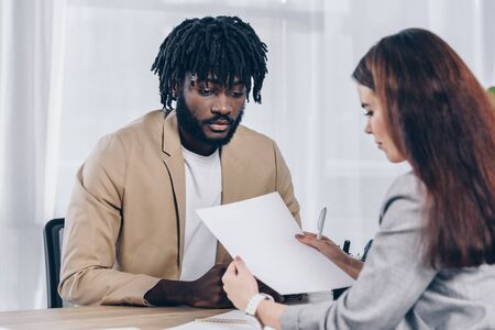 Selective focus of recruiter looking at paper near worried african american employee at job interview in officeの写真素材