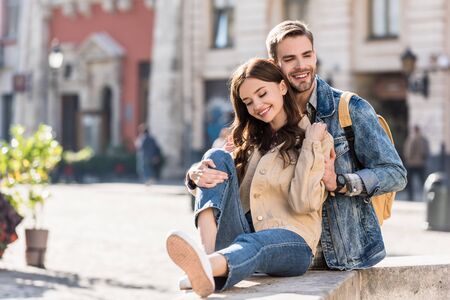 Selective focus of boyfriend hugging girl sitting on stone surface and smiling in cityの写真素材