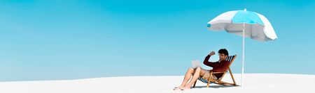 freelancer sitting with laptop in deck chair under umbrella on sandy beach against blue sky, panoramic shotの写真素材
