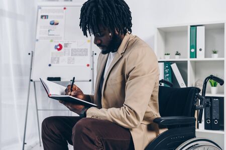 African american disabled employee on wheelchair writing in notebook in officeの写真素材