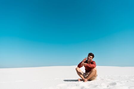 happy man on sandy beach talking on smartphone and looking at wristwatch against clear blue skyの写真素材