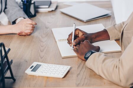 Cropped view of recruiter conducting job interview with employee and writing in notebook at table in...