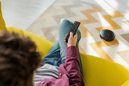 selective focus of man sitting on sofa and watching movie while robotic vacuum cleaner washing...