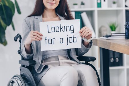 Cropped view of disabled employee smiling and holding placard with looking for a job lettering on...