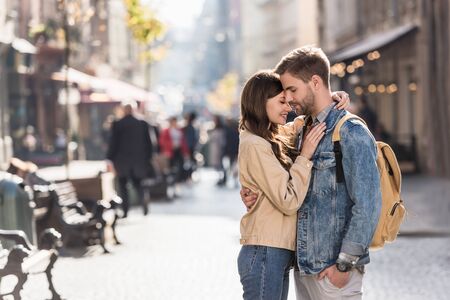 Boyfriend with hand in pocket and girlfriend hugging with closed eyes in city in Europeの写真素材