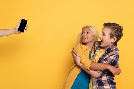 cropped view of woman holding smartphone and scared kids hugging and screaming on yellow background