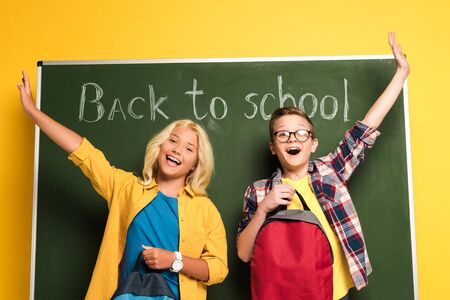 low angle view of smiling schoolkids with outstretched hands holding backpacks and standing near chalkboard with back to school letteringの写真素材
