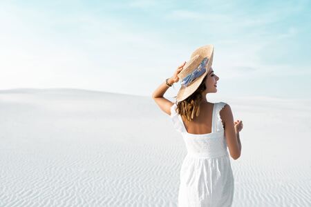 back view of beautiful girl in white dress and straw hat on sandy beach with blue skyの写真素材