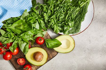 Top view of fabric near plate with greenery and cutting board with cherry tomatoes, basil leaves and avocado halves on greyの写真素材