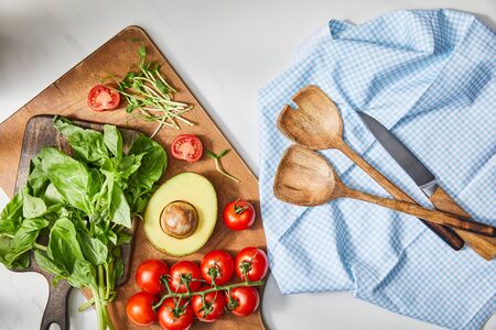 Top view of basil, cherry tomatoes, avocado half and microgreens on cutting boards near cloth with spatulas and knife on whiteの写真素材