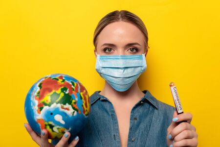 young woman in medical mask holding test tube with coronavirus blood sample and globe on yellow...