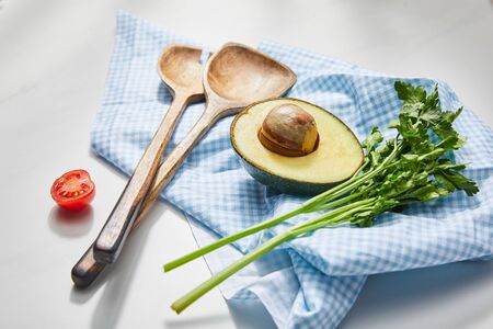 Selective focus of parsley, spatulas and avocado half on plaid cloth near cherry tomato on white...