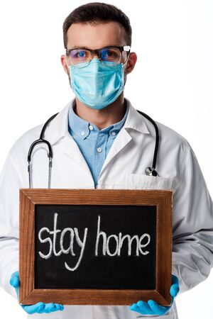 doctor in medical mask and white coat holding chalk board with stay home lettering isolated on whiteの写真素材