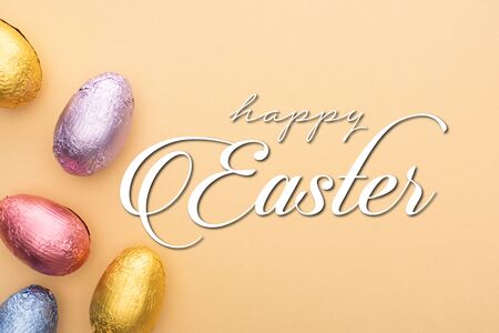Top view of chocolate Easter eggs in colorful foil on beige background with happy Easter...