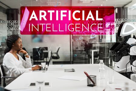 smiling african american businesswoman and robot sitting at desk in conference hall, Artificial Intelligence illustrationの写真素材