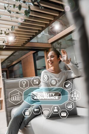 selective focus of happy asian businesswoman waving hand while sitting on couch near laptop, quality assurance illustrationの写真素材