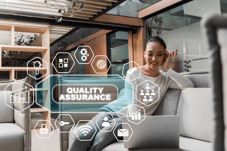 selective focus of smiling asian businesswoman sitting on couch and using laptop, quality assurance illustrationの写真素材