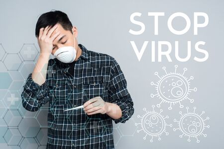 young asian man in respirator mask looking at thermometer and touching forehead on grey background, stop virus illustrationの写真素材