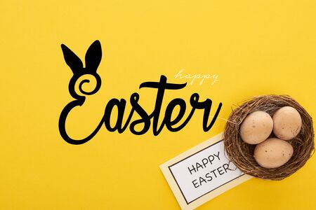 top view of greeting card with happy Easter lettering near chicken eggs in nest on yellow colorful backgroundの写真素材