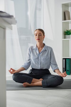 attractive businesswoman practicing yoga in lotus position with gyan mudra on mat in officeの写真素材