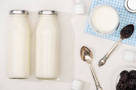 Top view of bottles and glass of yogurt on cloth, teaspoons, blackberries and containers with starter cultures on whiteの写真素材