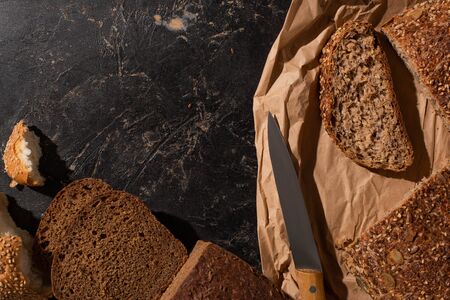 top view of cut whole grain bread on paper near knife on stone black surface