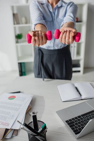 cropped view of businesswoman training with dumbbells at workplace with laptop and paperworkの写真素材