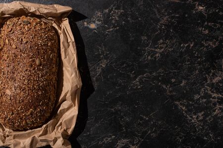 top view of fresh baked loaf of whole grain bread in paper on stone black surfaceの写真素材