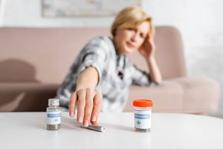 selective focus mature woman taking rolled joint with legal marijuana near bottles with medical cannabis letteringの写真素材