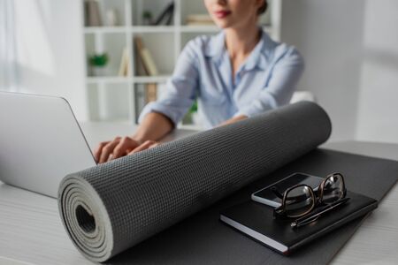 selective focus of young businesswoman working on laptop at workplace with yoga mat, notepad,...