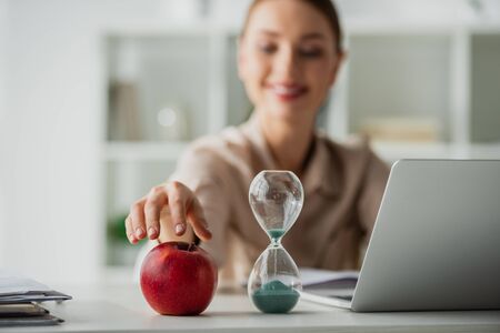 selective focus of positive beautiful businesswoman sitting at workplace with apple, sand clock and laptop in officeの写真素材