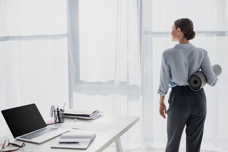 back view of businesswoman holding yoga mat at workspace with laptop and notepadの写真素材