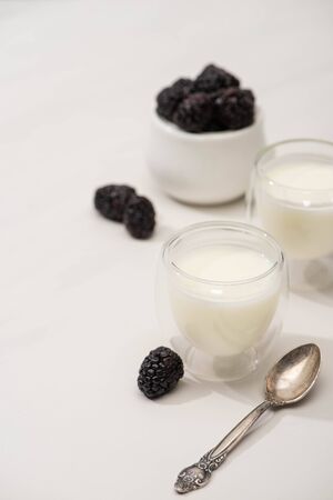 Selective focus of glasses of homemade yogurt, teaspoon and sugar bowl with blackberries on white backgroundの写真素材