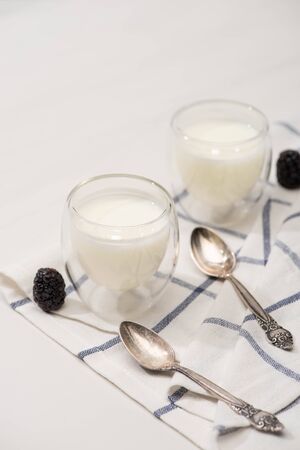 High angle view of glasses of homemade yogurt, teaspoons and blackberries on cloth on white backgroundの写真素材