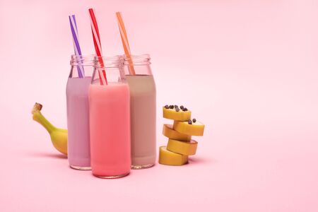 Bottles of berry, strawberry and chocolate milkshakes with drinking straws and cut banana with chocolate chips on pink backgroundの写真素材