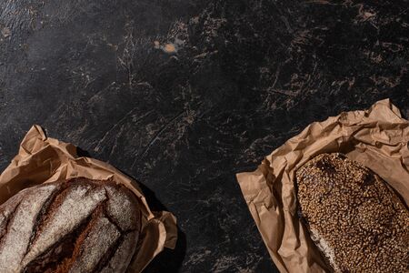 top view of fresh baked whole grain bread loaves in paper on stone black surface